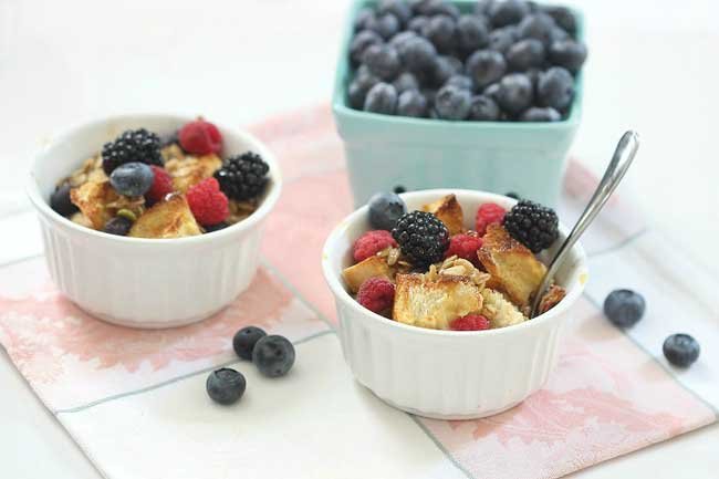 2 small white bowls of berry french toast casserole with a square container of blueberries and some blueberries scattered on the table