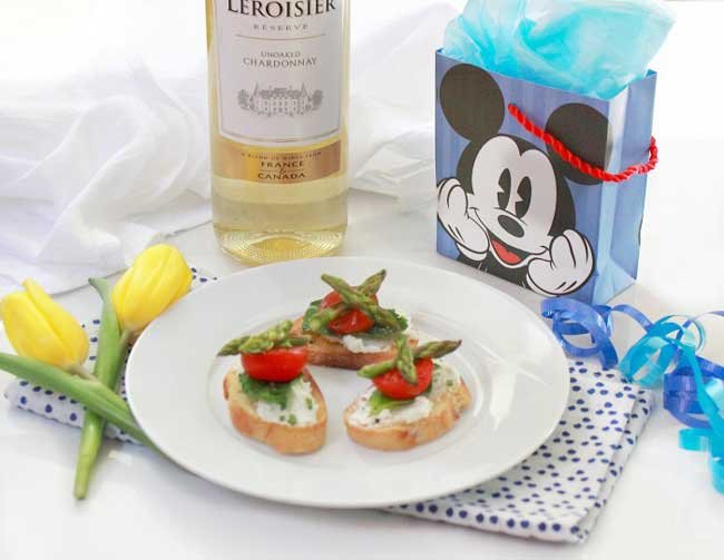 plate of 3 bruschetta's with mozzarella, asparagus and tomato with a bottle of white wine, 2 yellow flowers and a gift bag