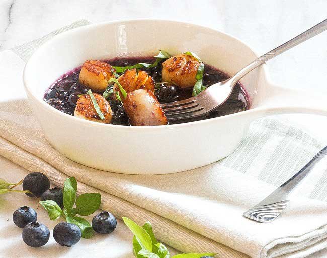 a bowl of pan seared sea scallops in blueberry sauce on a white table cloth with a few blueberries scattered on the table